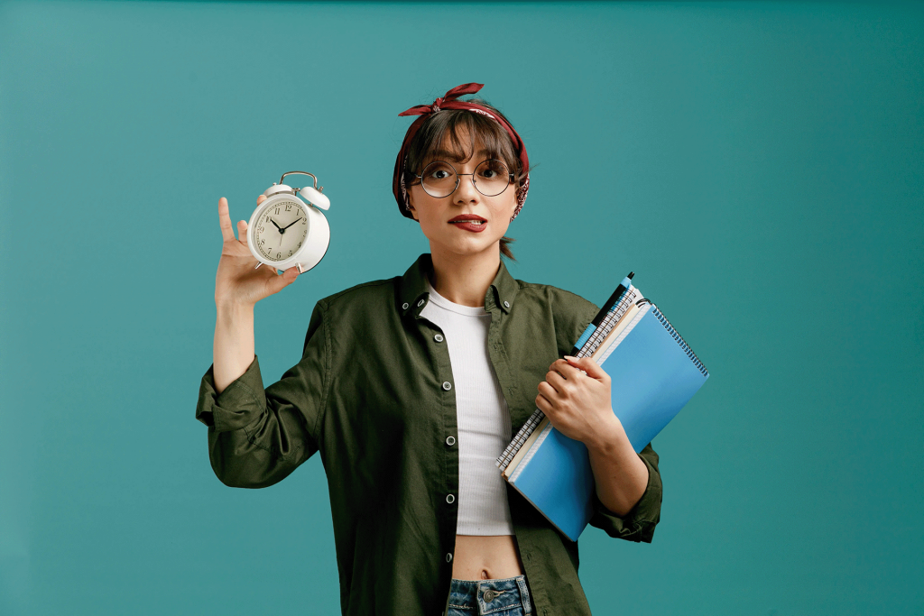 anxious-young-student-girl-wearing-bandana-glasses-holding-large-note-pads-with-pen-looking-camera-biting-lip-showing-alarm-clock-isolated-blue-background